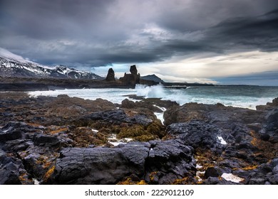 Waves crash onto the shoreline in front of Longdrangar, a pair of basalt rock outcrops on the Snaefellsnes Peninsula, Iceland. Volcanic rock and seaweed foreground with snow capped mountains beyond. - Shutterstock ID 2229012109