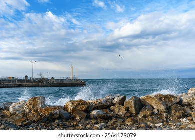 Waves Crashing on Rocks, Cloudy Sky, Rocky Coastline with Jetty and Seagulls or gulls. Yellow navigation light post or Lighthouse on the concrete pier. Waterfront View with Seabirds - Powered by Shutterstock