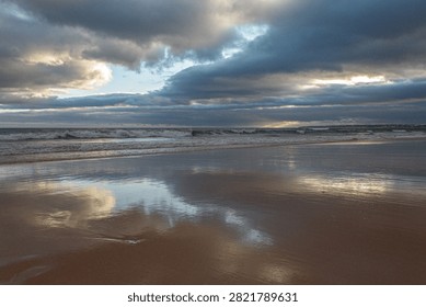 Waves gently lap against the sandy shore at Praia da Gale as the sun begins to set, casting a golden glow over the landscape. Dark clouds subtly shift in the sky, with hints of blue peeking through, creating a dynamic backdrop. The wet sand mirrors the stunning colors above, enhancing the serene beauty of the scene. Seagulls can be seen in the distance, gliding effortlessly through the cool evening air.
