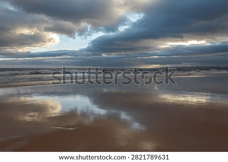Waves gently lap against the sandy shore at Praia da Gale as the sun begins to set, casting a golden glow over the landscape. Dark clouds subtly shift in the sky, with hints of blue peeking through, creating a dynamic backdrop. The wet sand mirrors the stunning colors above, enhancing the serene beauty of the scene. Seagulls can be seen in the distance, gliding effortlessly through the cool evening air.