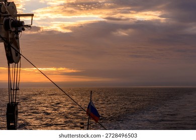 Waves gently ripple as the sun sets beneath a colorful sky. A ship's flag flutters in the breeze, marking time on the open ocean during twilight.
