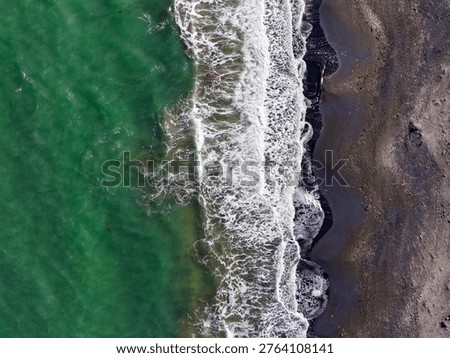 Waves move toward the shore creating foam as they meet the rocky beach. The water appears green in color under bright sunlight. The sandy and rocky textures are clear.