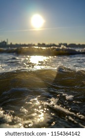 Waves Rapidly Approaching A Hudson River Bank With New York City And Sun In Soft Focus In The Background.