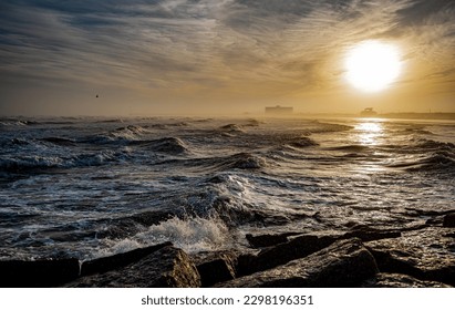 Waves roll onto a beach as the sun is low in the sky, in ominous dramatic seascape landscape.