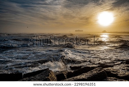 Waves roll onto a beach as the sun is low in the sky, in ominous dramatic seascape landscape.