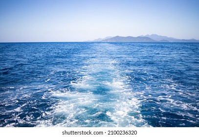 Waves From Slowly Moving Boat Or Ship. View Of A Green Island On The Horizon. No People. Clear Sky.