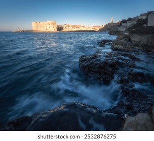 Waves splash gently against rugged rocks by a sunlit, historic coastal fortress under a clear blue sky. - Powered by Shutterstock