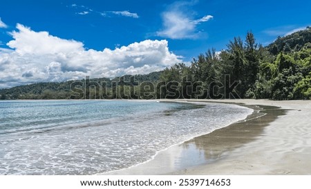 The waves of the turquoise ocean spread over the sandy beach. Delicate foam and reflection on wet sand. A hill, green vegetation against the blue sky and clouds. Malaysia. Borneo. Kota Kinabalu.