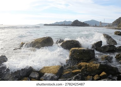 Waves vigorously splash on rugged moss covered rocks along the shoreline with a busy port visible in the distant background