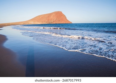 Waves washing ashore on volcanic sand with view of Montaña Roja mountain in Tenerife Spain symbol of travel freedom and natural beauty