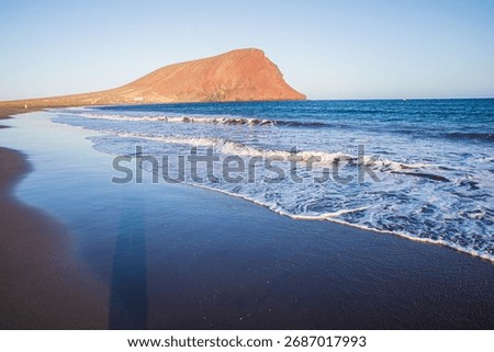 Waves washing ashore on volcanic sand with view of Montaña Roja mountain in Tenerife Spain symbol of travel freedom and natural beauty