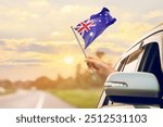 Waving the Australia flag against the sunrise or sunset from a car driving along a country road. Holding the Australia flag, traveling by car, on a weekend trip.