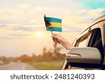 Waving the Bahamas flag against the sunrise or sunset from a car driving along a country road. Holding the Bahamas flag, traveling by car, on a weekend trip.