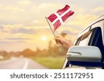 Waving the Denmark flag against the sunrise or sunset from a car driving along a country road. Holding the Denmark flag, traveling by car, on a weekend trip.