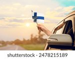Waving the Finland flag against the sunrise or sunset from a car driving along a country road. Holding the Finland flag, traveling by car, on a weekend trip.