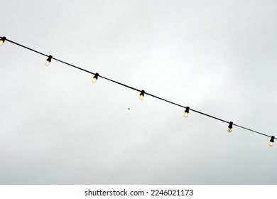 Waving hanging lighting garlands of 7 electric light bulbs on a black electric cable strand in the foreground of a gray rainy sky in winter holidays on the Belgian coast. One light is stuck - Powered by Shutterstock