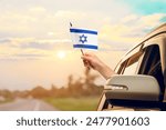 Waving the Israel flag against the sunrise or sunset from a car driving along a country road. Holding the Israel flag, traveling by car, on a weekend trip.