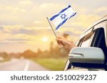 Waving the Israel flag against the sunrise or sunset from a car driving along a country road. Holding the Israel flag, traveling by car, on a weekend trip.