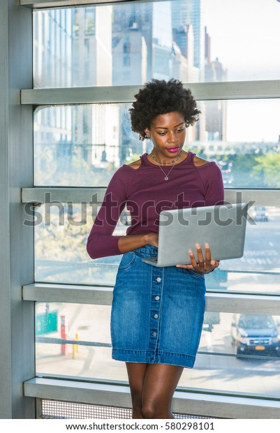 Way to\
success. African American college student with short afro hairstyle\
stands against glass wall on campus in New York, works on laptop\
computer. Street, cars on\
background.\
