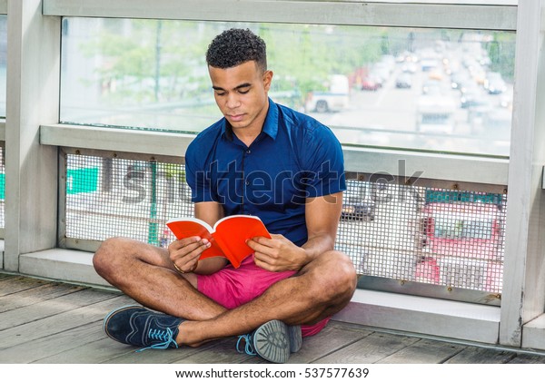 Way to Success. African American male college\
student sitting cross legged on floor against glass wall on campus\
in New York, reading red book. Street, many cars on background.\
Color filtered effect\
