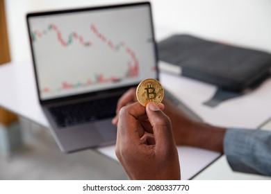 We Can See The Hand Of An African American Who Is Holding A Physical Bitcoin He Is In Front Of His Laptop Staring At A Graphic That Goes Down