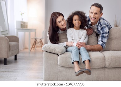 We cherish you. Nice exuberant curly-haired girl smiling and sitting on the couch and her parents standing behind her - Shutterstock ID 1040777785
