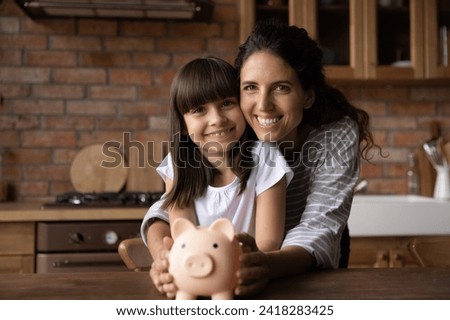 We choose wise economy. Portrait of happy little latin girl and single mom sit at kitchen table cuddle look at camera hold toy piggy bank. Thrifty mum parent and preteen kid make deposit save up money