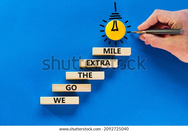 We go the\
extra mile symbol. Wooden blocks with words We go the extra mile.\
Light bulb icon. Businessman hand, pen. Blue background, copy\
space. Business, we go the extra mile\
concept.