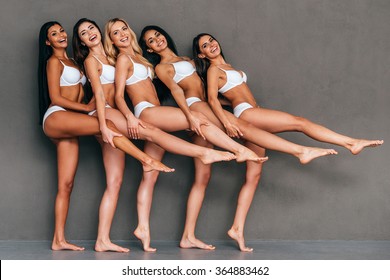 We Got Perfect Legs! Group Of Five Beautiful Women In Lingerie Stretching Out Their Legs And Smiling While Standing Against Grey Background