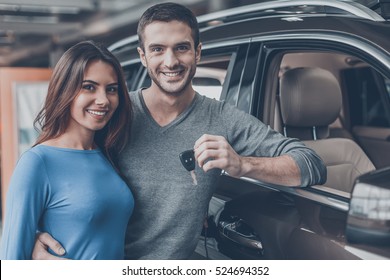 We have made the right choice. Handsome young men standing near the car at the dealership hugging his girlfriend and holding a key