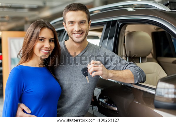 We have made the right choice. Handsome young men\
standing near the car at the dealership hugging his girlfriend and\
holding a key
