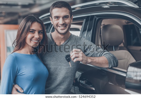 We have made the right choice. Handsome young men\
standing near the car at the dealership hugging his girlfriend and\
holding a key