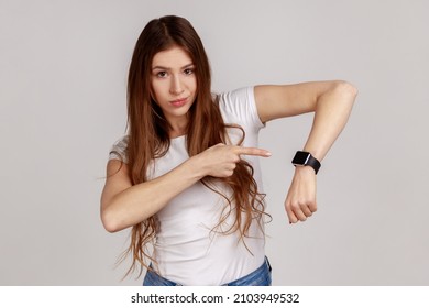 We have no time. Portrait of serious boss woman with dark hair pointing her finger at the smartwatch and getting angry, wearing white T-shirt. Indoor studio shot isolated on gray background.