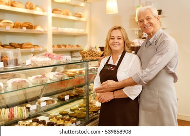 We keep delicious goods baked. Portrait of a senior loving couple running their bakery together posing near the showcase full of delicious desserts welcoming to their shop smiling to the camera - Powered by Shutterstock