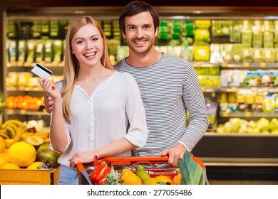 We Like Shopping Together. Cheerful Young Couple Smiling At Camera And Showing A Credit Card While Standing In A Food Store