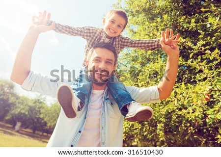 We like spending time together! Low angle view of happy little boy stretching out hands while his father carrying him on shoulders