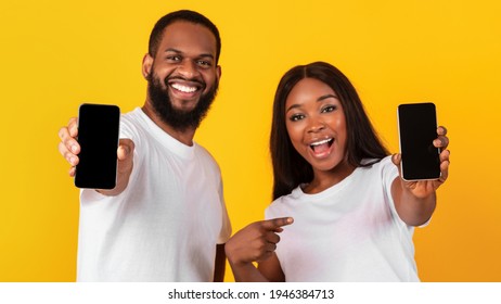 We Like This App. Cheerful African American Couple Holding And Presenting Their Two Mobile Phones With Black Empty Screen In Hands, Lady Pointing Fingers At Gadget With Blank Space For Mock Up