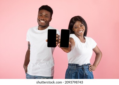 We Like This App. Cheerful African American Couple Holding And Presenting Their Two Mobile Phones With Black Empty Screen In Hands, Showing 2 Gadgets With Blank Space For Mock Up, Pink Studio Wall