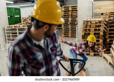 We lose control, let's get loud. A man and a woman are playing in the warehouse next to the wooden pallets. He is pulling a hand pallet truck while she maintains her body balance. Forever children - Powered by Shutterstock