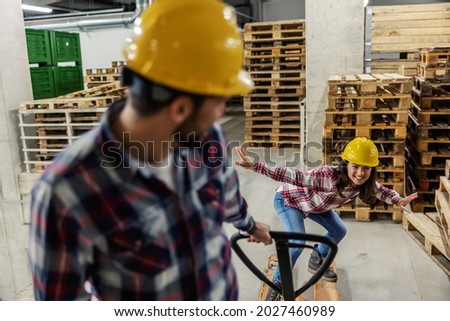 We lose control, let's get loud. A man and a woman are playing in the warehouse next to the wooden pallets. He is pulling a hand pallet truck while she maintains her body balance. Forever children