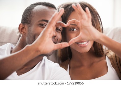 We Love Each Other! Beautiful Young African Couple Sitting Close To Each Other And Looking Through A Heart Shape Made With Their Fingers