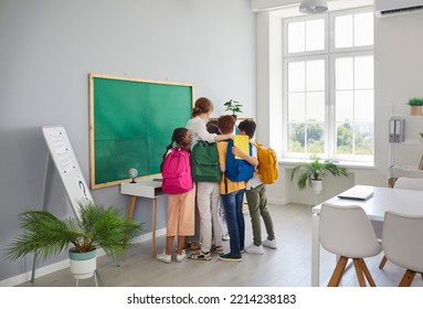 We Love Our Teacher. Happy Elementary School Kids Hugging Their Female Teacher Thanking Her Or Celebrating Back To School. Schoolchildren With Backpacks On Shoulders Hug Woman In School Classroom.