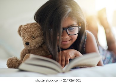 We love storytime, anytime. Shot of a cute little girl reading a book in her bedroom with her teddybear by her side.