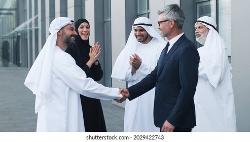 We made a deal. Full length view of successful two business partners shaking hands. They are standing at the street near their colleagues. People are applauded at the background
