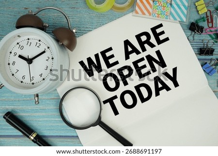 We Are Open Today text on a paper surrounded by office supplies, a magnifying glass, and a clock on a blue wooden surface
