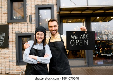 We are OPEN. Two multiracial friendly waiters wearing uniform stand outside a restaurant, cafe or bar, showing signboard OPEN, with pleasured smiling, welcome. Teamwork concept