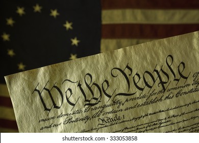 We The People, The Opening Words Of The Preamble To The Constitution Of The USA, Is Prominent In This Image. The Parchment Document Is In Front Of On An Aged Grungy Colonial Era American Flag. 