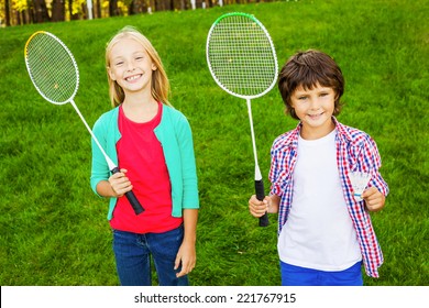 We are ready to play! Two cute little children holding badminton rackets and smiling while standing on green grass together