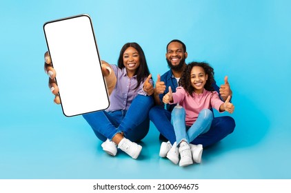 We Recommend. Smiling black family holding cell phone with white blank device screen in hand, showing thumb up gesture, blue studio. Gadget with empty free space for mock up template, banner isolated - Powered by Shutterstock