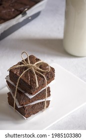 We see a stack of three brownies wrapped and served on a white plate  in the background, we can see a bottle of milk 